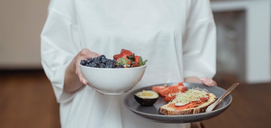 Person holding a bowl of fresh berries and a plate with avocado toast, displaying a healthy breakfast spread.